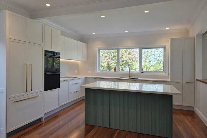 A finished kitchen with warm off-white shaker cabinets, a sage green fluted island, pale stone benchtops and a wide window looking onto the garden