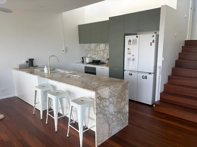 A finished kitchen with muted green pantry cabinets, white lower drawers, a veined marble waterfall island and breakfast bar seating beside a timber staircase