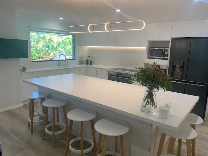 A bright white kitchen with a large central island, matte black appliances, light timber floors and a wide window over the sink